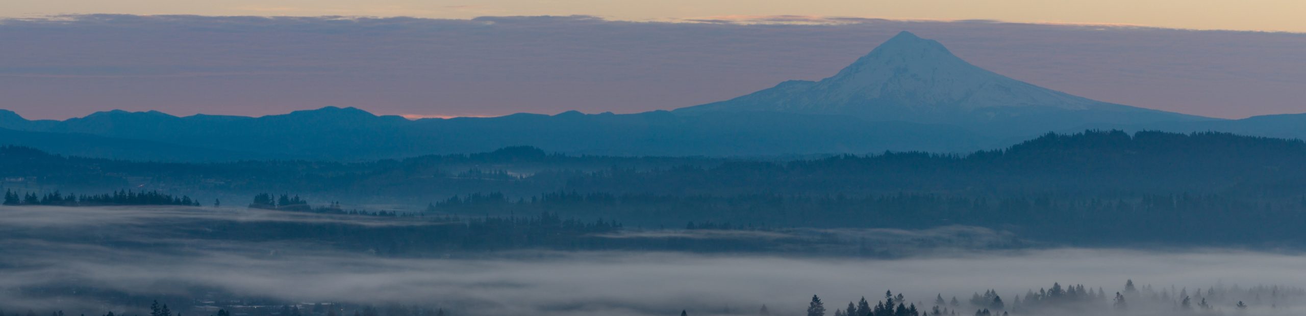 Wide landscape at dawn showing a snow-capped mountain peak rising above misty blue hills and forest.